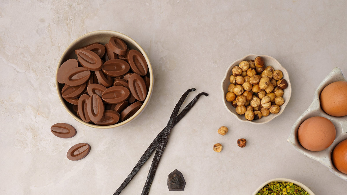 Valrhona baking fèves in a bowl beside vanilla beans, hazelnuts, eggs and pistachios on a stone counter