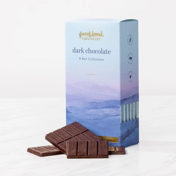 A blue watercolor mountain box of French Broad dark chocolate 6 Bar Collection behind unwrapped chocolate pieces.