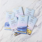 Six French Broad dark chocolate bars fanned on white marble. The light blue wrappers feature watercolor illustrations.