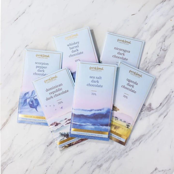 Six French Broad dark chocolate bars fanned on white marble. The light blue wrappers feature watercolor illustrations.