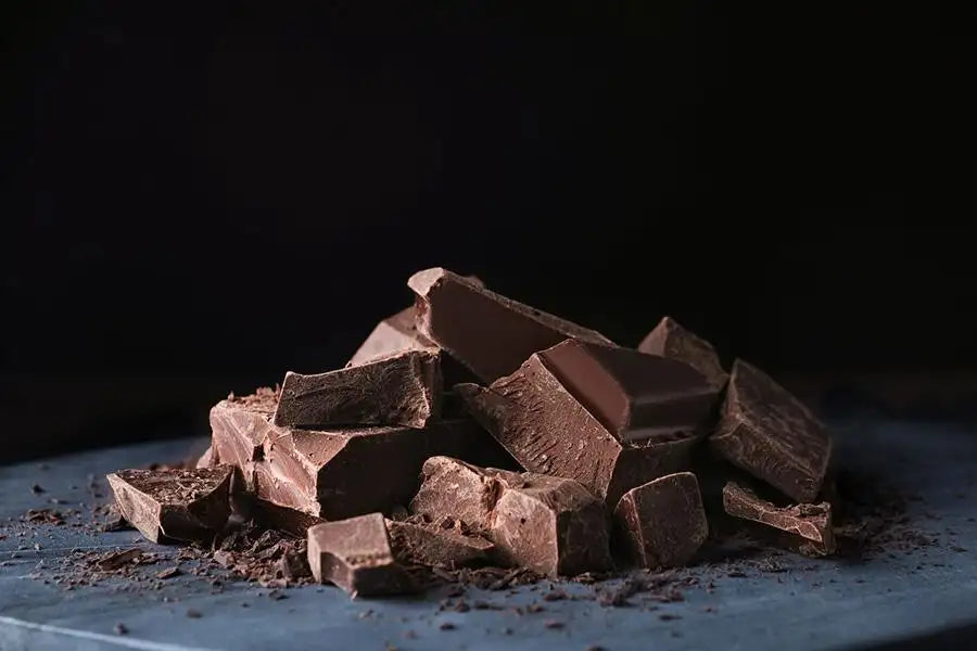 A pile of dark chocolate chunks with a rich, glossy surface and fine cocoa powder scattered around them.