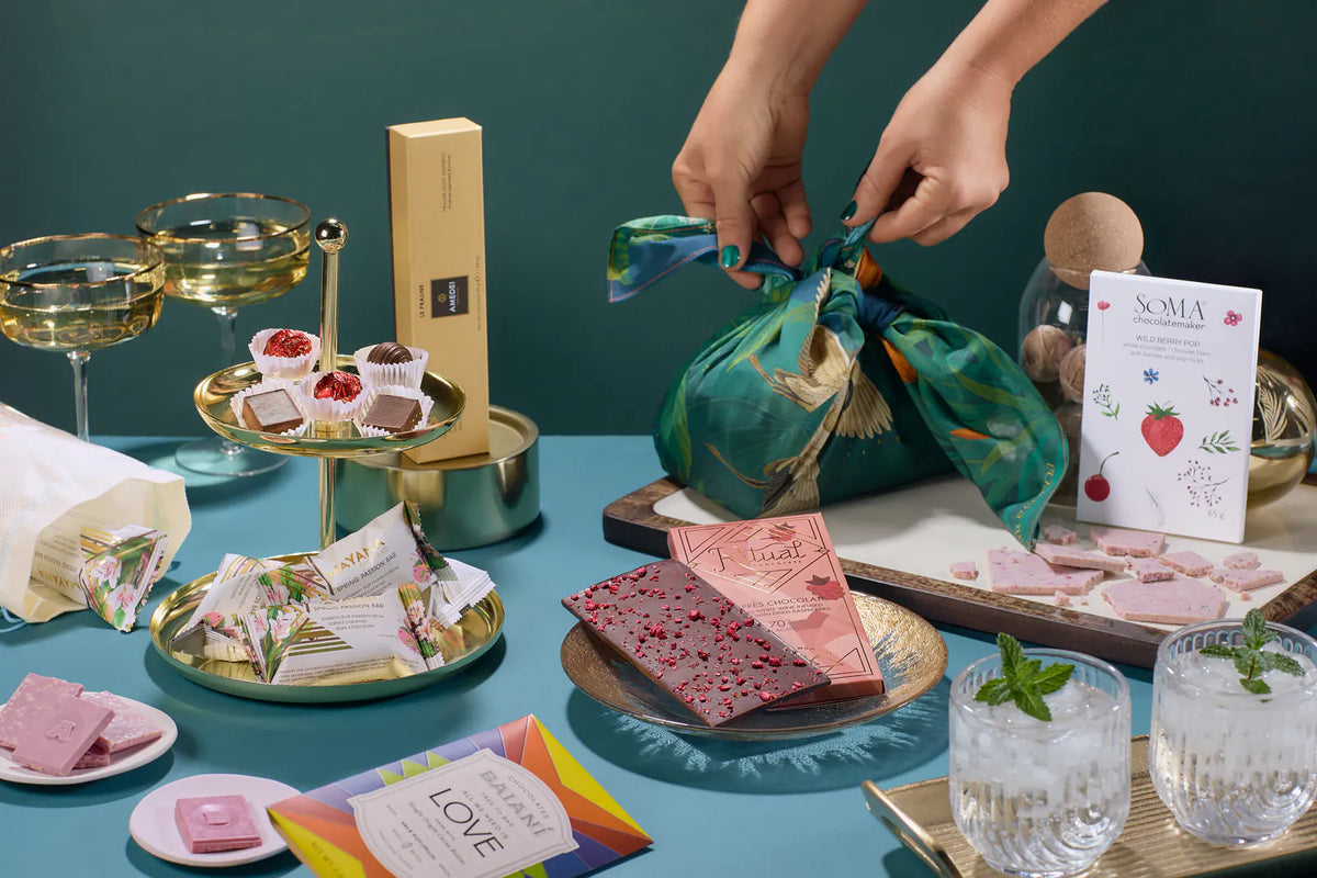 Hands tie Bar & Cocoa's teal botanical furoshiki cloth around a chocolate gift, surrounded by craft chocolate and glassware on a teal table.