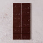 A dark chocolate bar with the word firetree embossed on alternating squares, resting on a textured, light-colored surface.