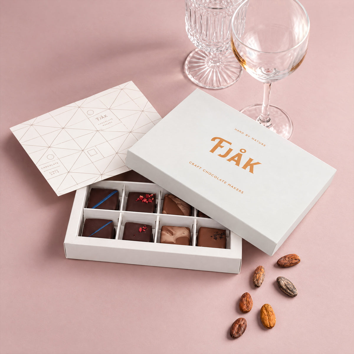 Open Fjåk box of 12 chocolate-coated mixed caramels with cacao beans, on a pink background.