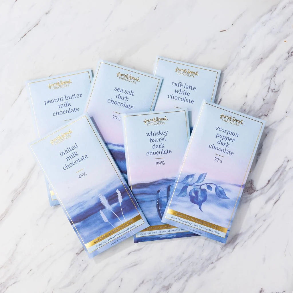Six French Broad Chocolate bars in light blue illustrated wrappers fanned out on a white marble surface.