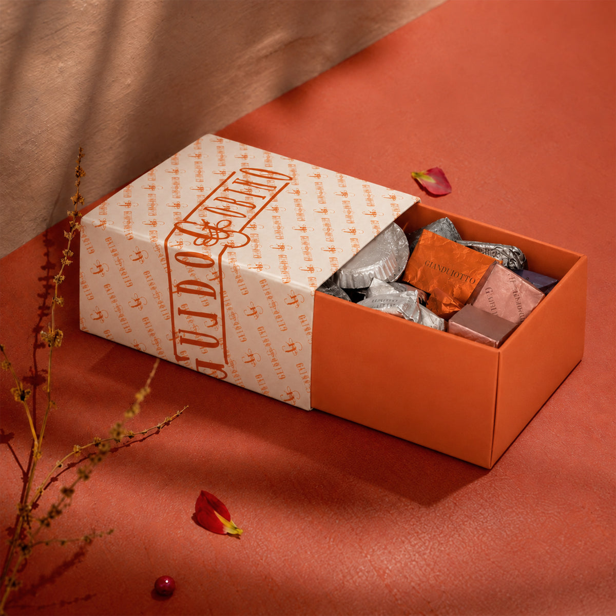 Open Guido Gobino gift box filled with foil-wrapped gianduiotti and chocolates, on an orange surface with dried branches and red petals.