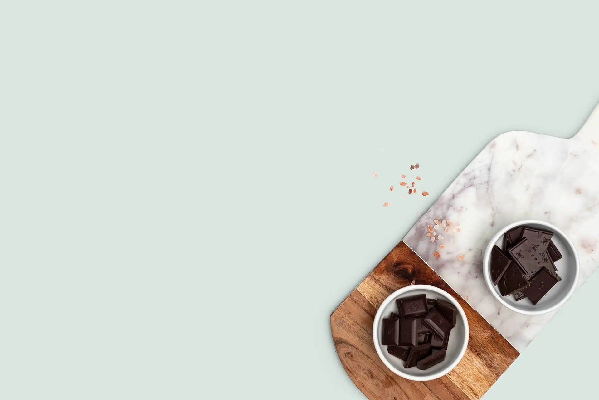 Dark chocolate squares in white ceramic bowls on a wooden cutting board and marble surface.