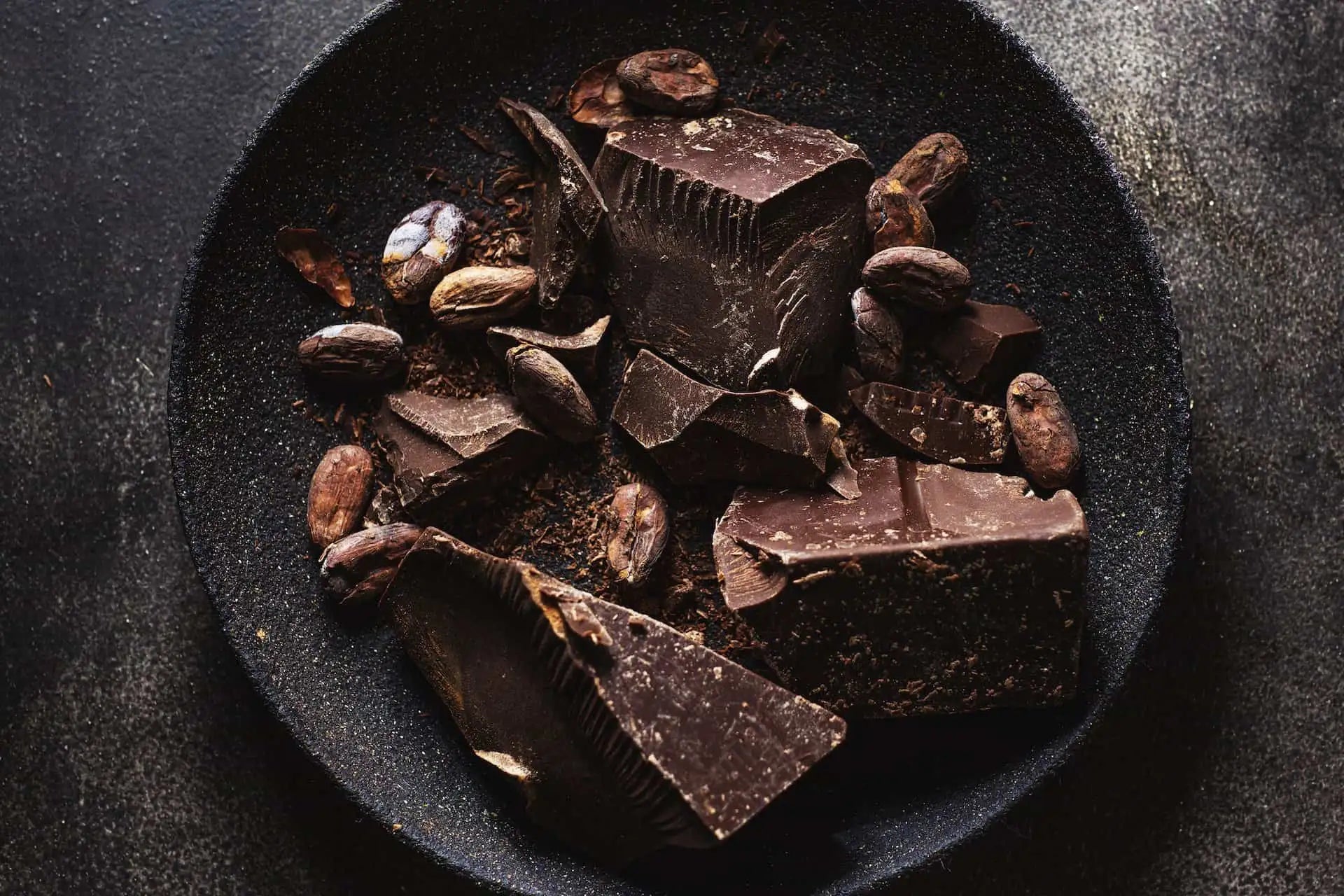 Dark chocolate chunks and cocoa beans scattered on a black plate.