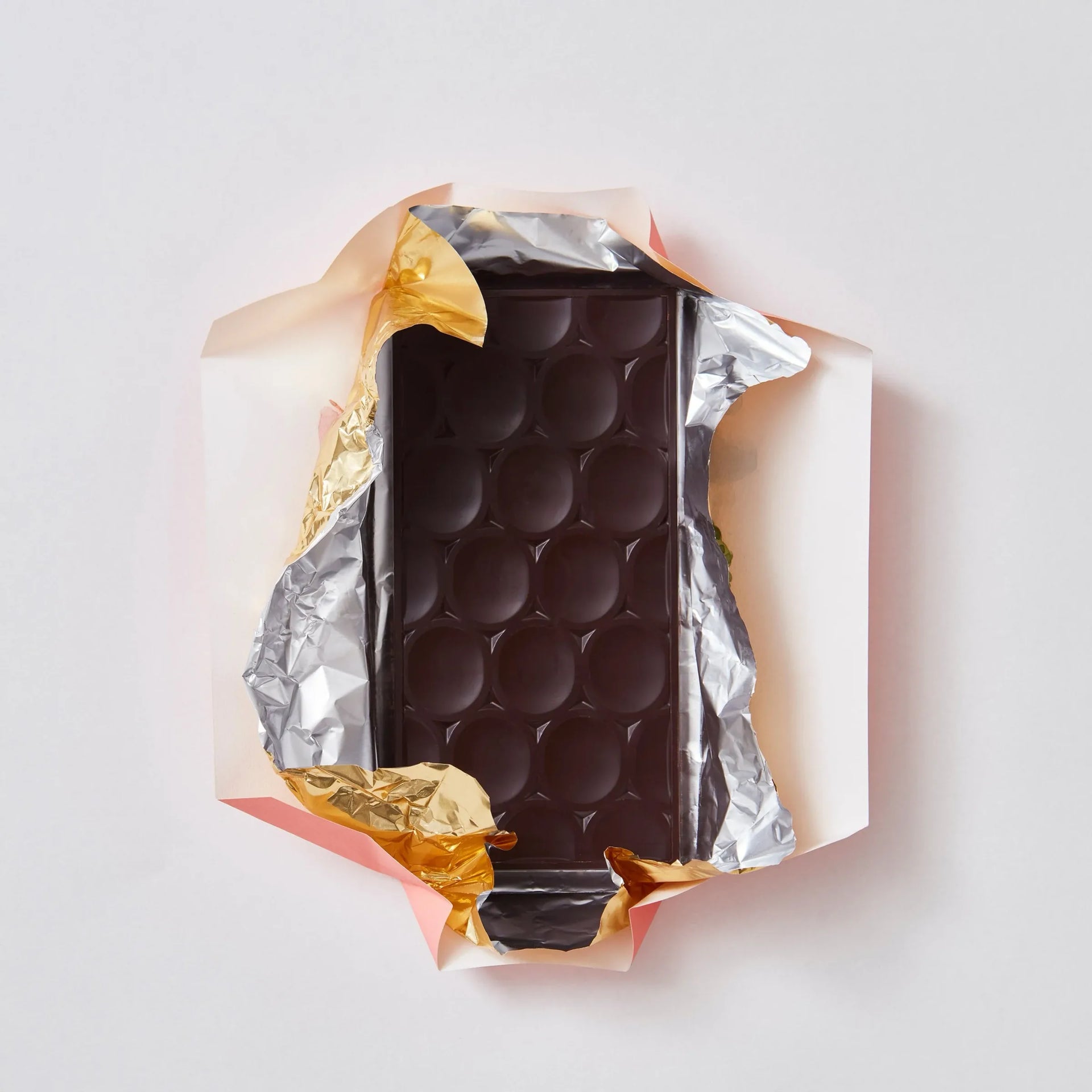 Dark chocolate bar with oval-relief pattern half unwrapped from gold and silver foil on a pale background