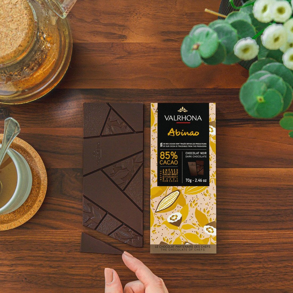 Hand holding a Valrhona Abinao 85% dark chocolate bar over broken squares on a wooden table