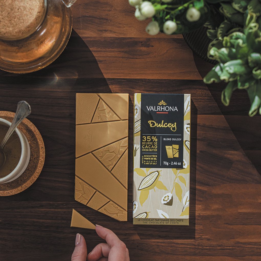 Hand holding a Valrhona Dulcey 35% blond chocolate bar over golden squares on a wooden table
