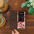Hand holding a Valrhona Guanaja 70% dark chocolate bar over broken squares on a wooden table