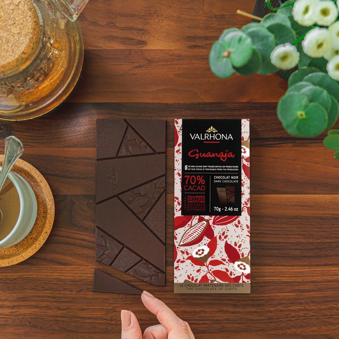 Hand holding a Valrhona Guanaja 70% dark chocolate bar over broken squares on a wooden table