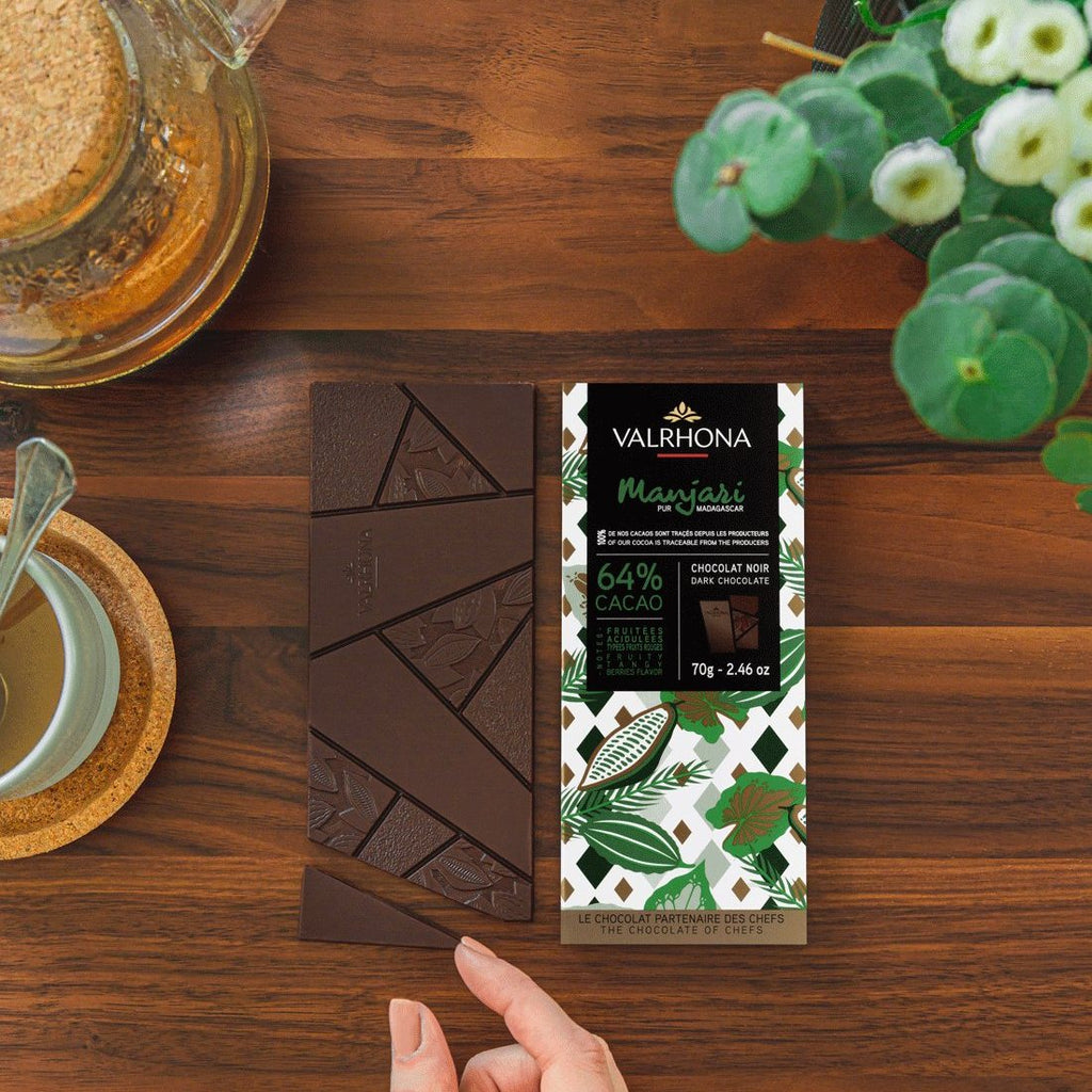 Hand holding a Valrhona Manjari 64% dark chocolate bar over broken squares on a wooden table