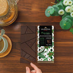 Hand holding a Valrhona Manjari 64% dark chocolate bar over broken squares on a wooden table