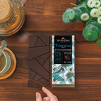 Hand holding a Valrhona Tulakalum 75% dark chocolate bar over broken squares on a wooden table
