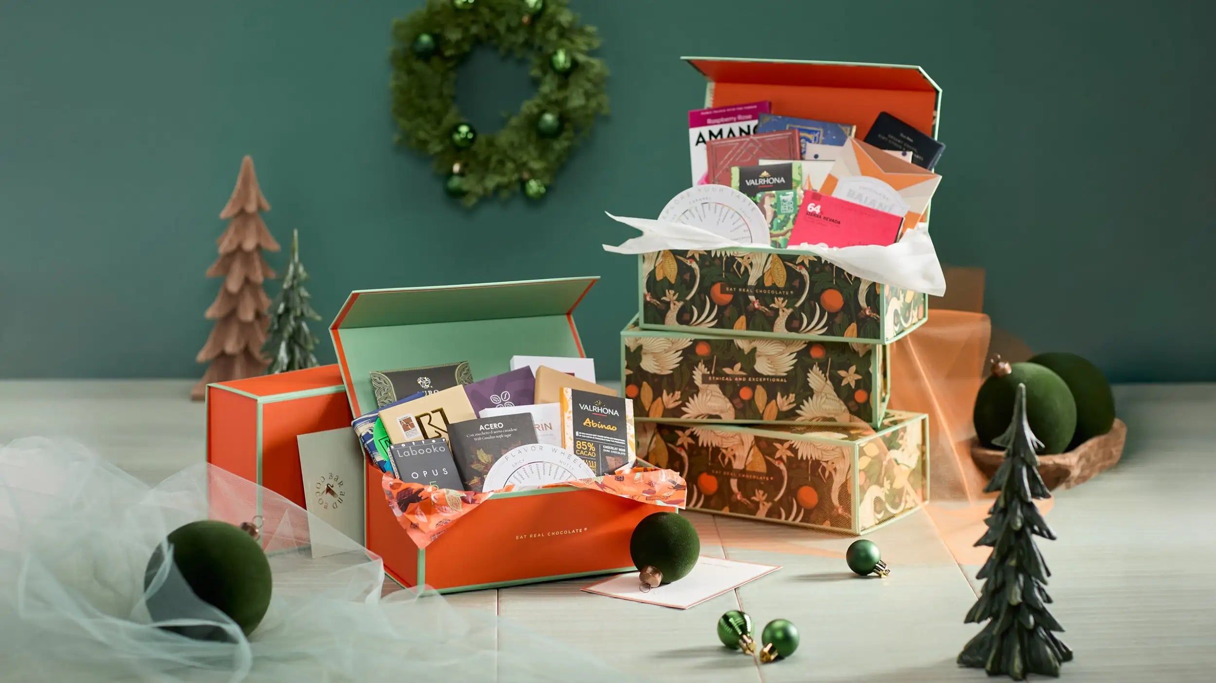 Open orange gift box with festive green and gold patterned interior, filled with assorted luxury items including chocolates, cards, and branded packaging.
