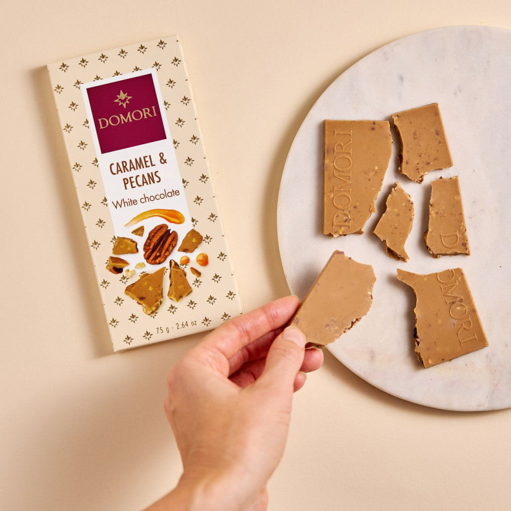 Hand holding broken Domori White Chocolate w/ Caramel & Pecans bar with packaging