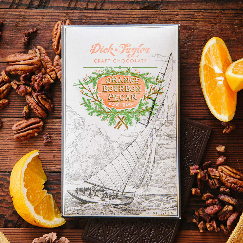 White Dick Taylor Orange Bourbon Pecan 65% Dark box with a sailboat drawing, surrounded by pecans and oranges on wood.